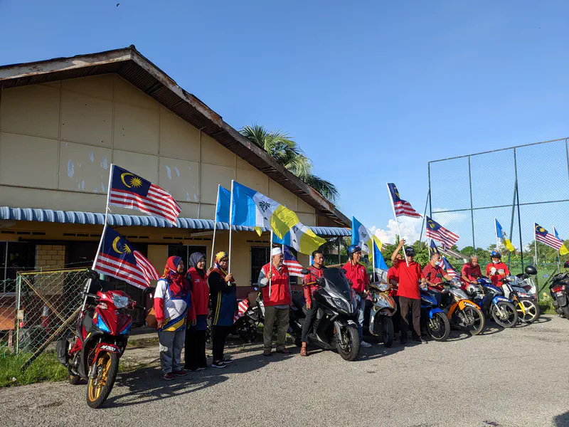 Side view of a group of people with motorcycles holding Malaysian and state flags, gathered beside a building with a fence.