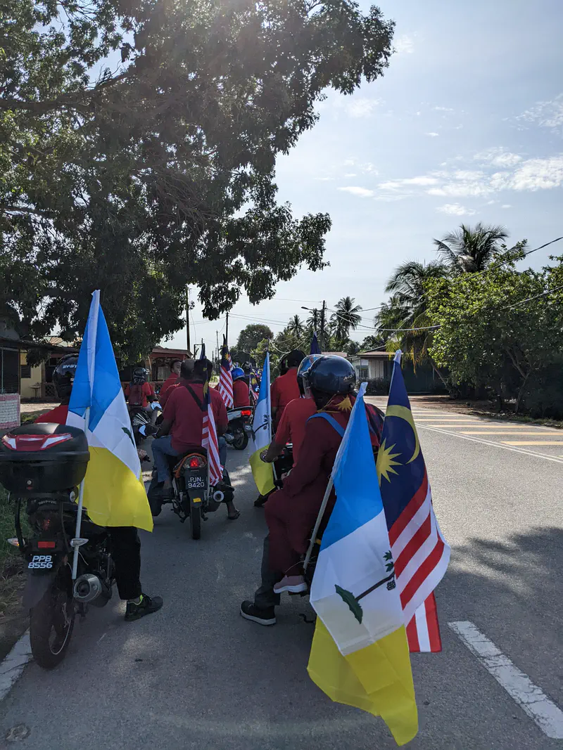 Group of motorcyclists wearing red shirts carrying Malaysian and state flags.