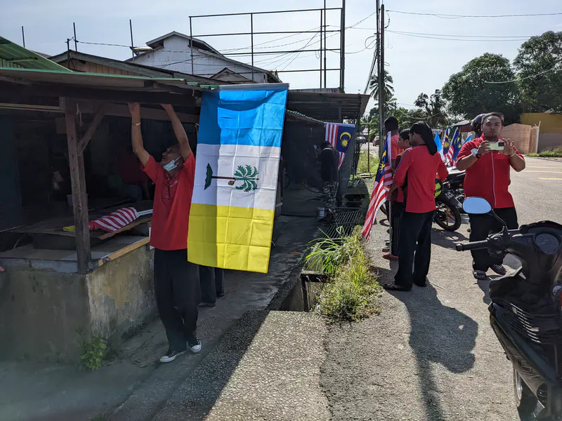Man in a red shirt hanging a Penang state flag outside a small building while others with Malaysian flags look on.