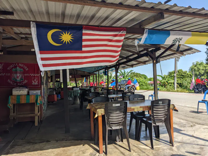 Open-air eatery with Malaysian and state flags hanging, motorcycles passing by outside.