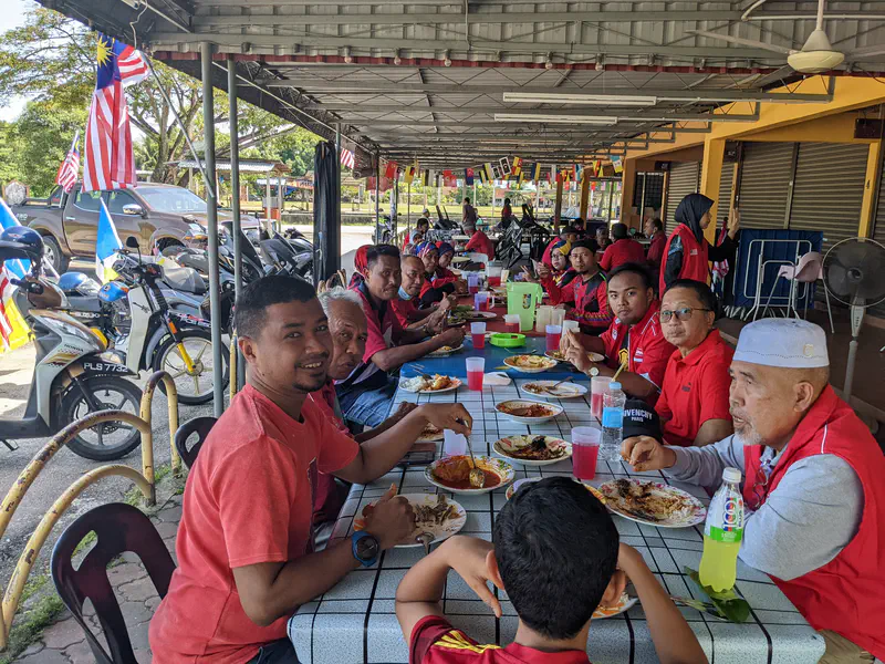 Group of people in red shirts sharing a meal at long tables under a covered area.