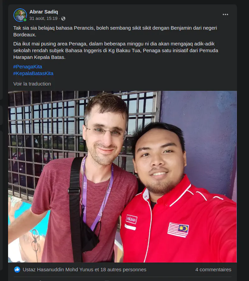 Two men taking a selfie together, one wearing a red shirt with the Malaysian flag and the other in a maroon shirt with glasses.