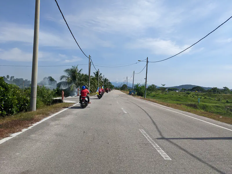 Group of motorcyclists riding along a paved road lined with power poles and greenery.