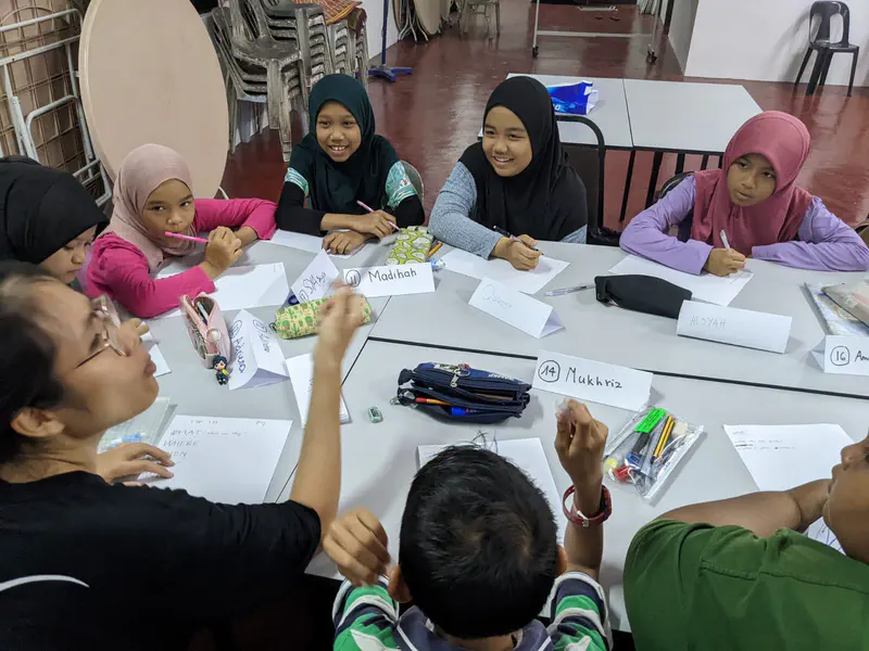 Children sitting around a table writing on paper with name tags in a classroom activity.