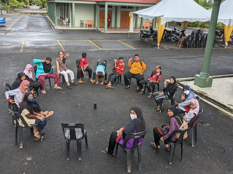 Group of children sitting in a large circle on chairs outdoors, smiling and waving.