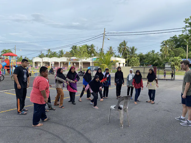 Children and a teacher playing a group game outdoors near a playground and road.