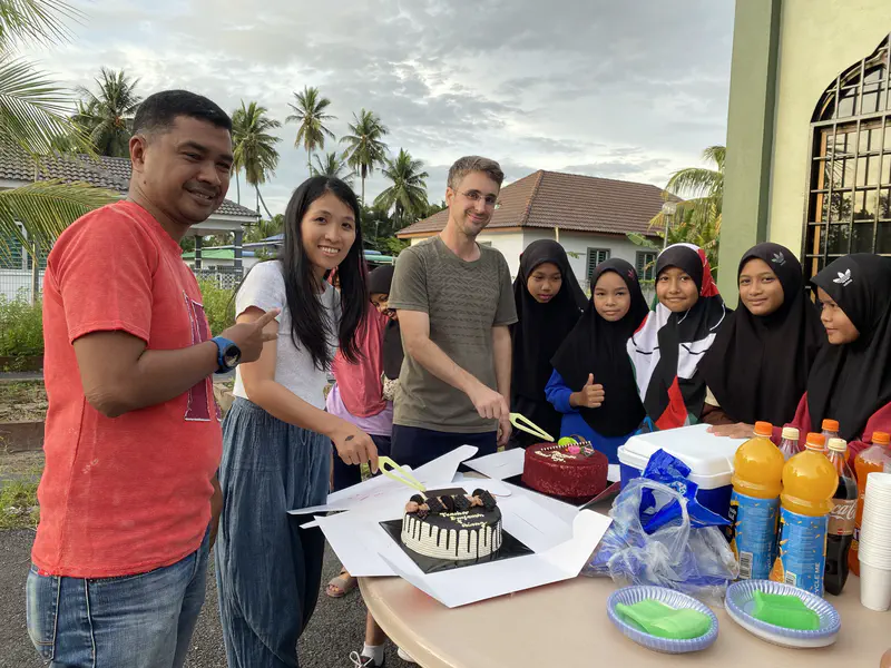 Group of people cutting cakes at an outdoor celebration with drinks and food on the table.