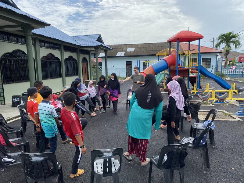 Children and a teacher playing a group game outdoors near a playground.