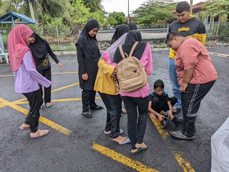 Group of children gathered around a boy sitting on the ground adjusting his sandal.