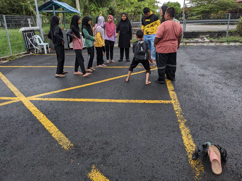 Children standing in a circle outdoors while a boy in the center prepares to play a game.