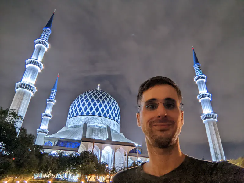 Person taking a selfie at night in front of a mosque with blue domes and tall minarets.