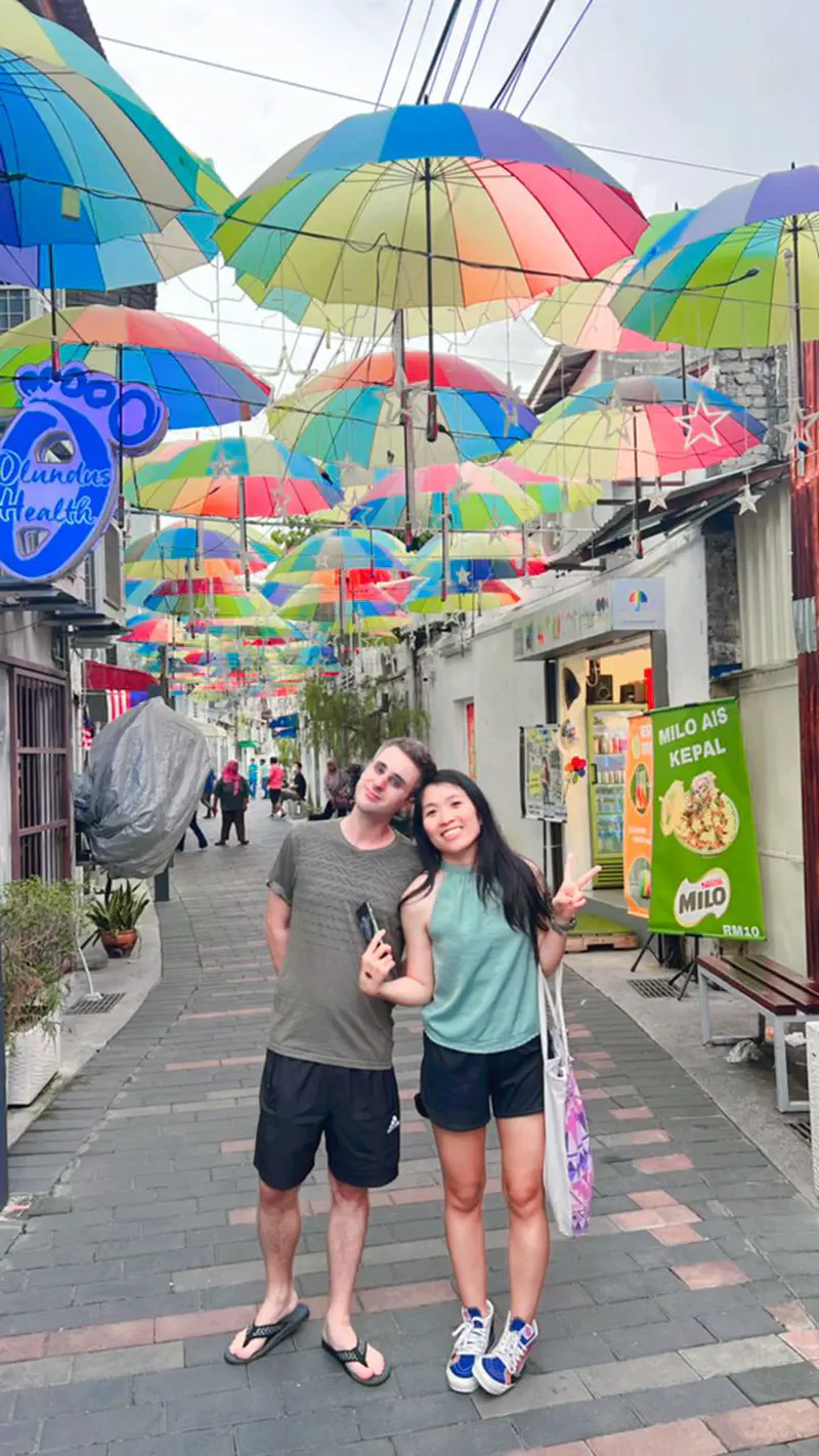 Two people posing under a street decorated with hanging colorful umbrellas.