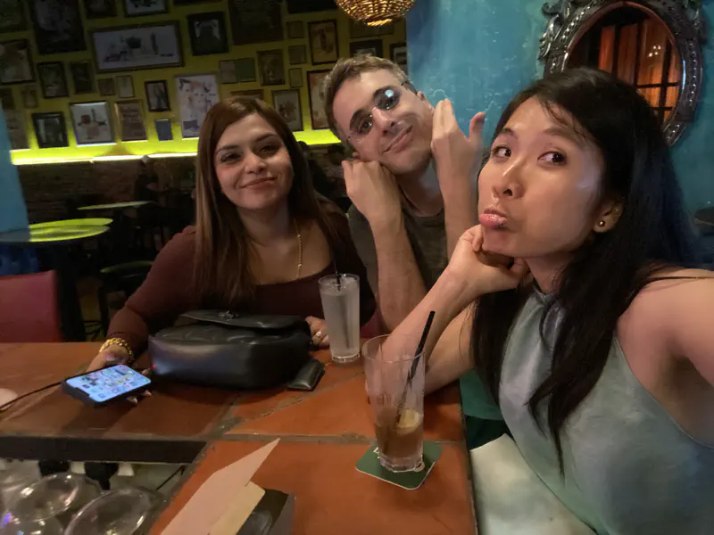 Three friends sitting at a table in a colorful cafe, smiling and posing for a selfie.