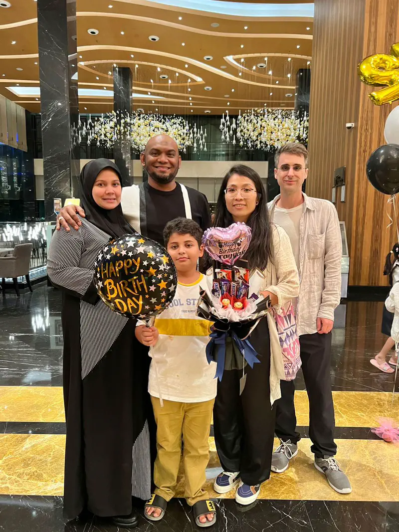 Two women, a man, and a boy posing together in a hotel lobby holding birthday balloons and a gift bouquet.