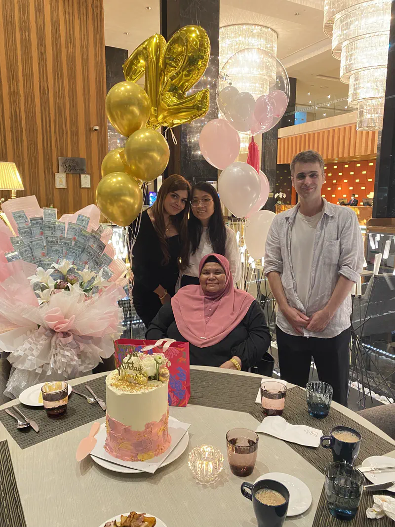 Three people standing behind an elderly woman at a birthday celebration with balloons and cake.