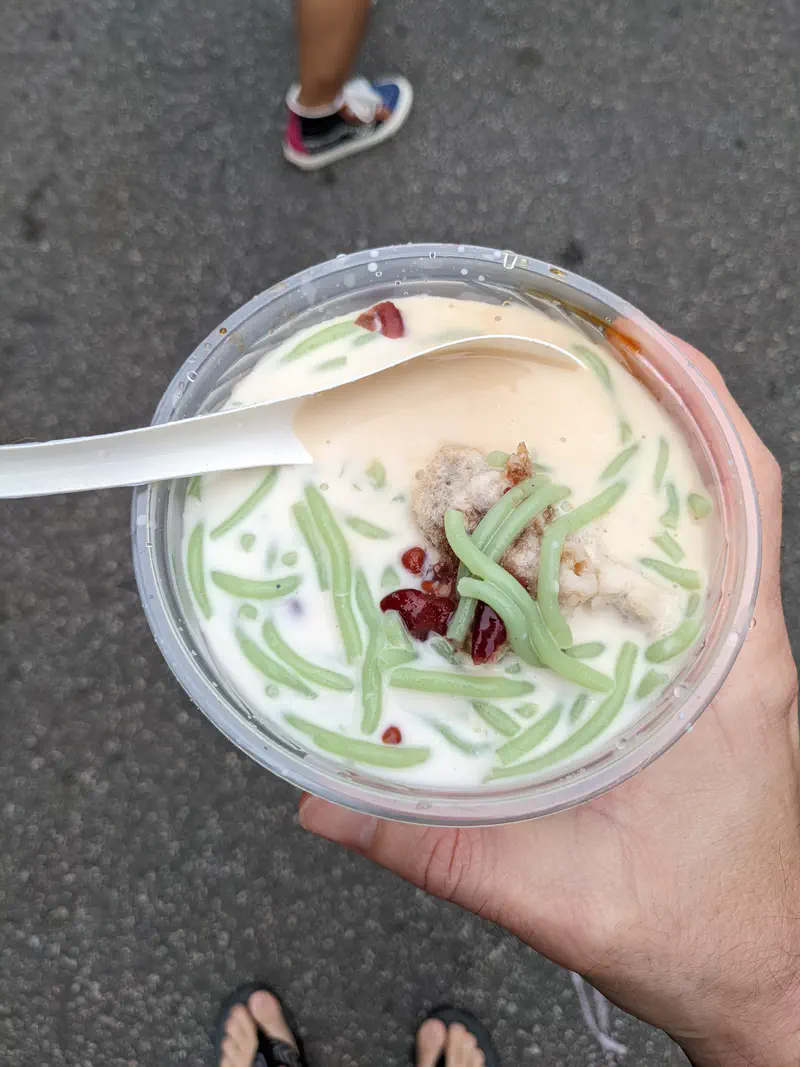 Hand holding a bowl of chendul dessert with green jelly, beans, and shaved ice in coconut milk.