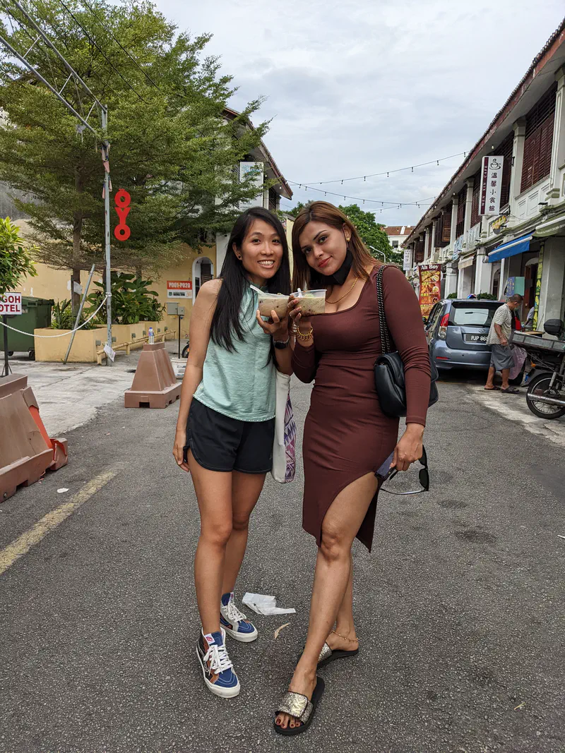 Two women standing on a street holding bowls of food and smiling at the camera.