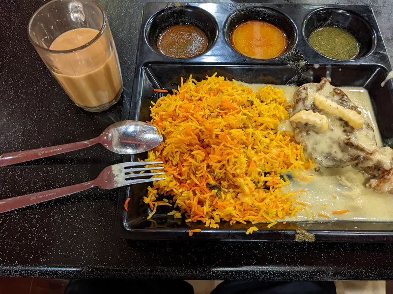 Plate of biryani rice with chicken in white sauce, three dipping sauces, and a glass of milk tea.
