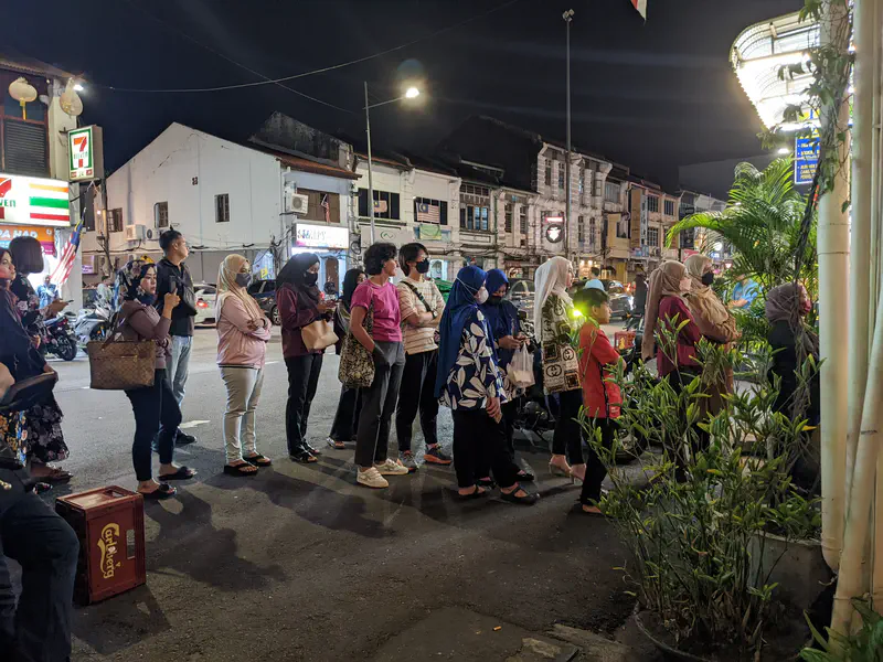 People standing in line outside at night along a street with shops and a 7-Eleven store.