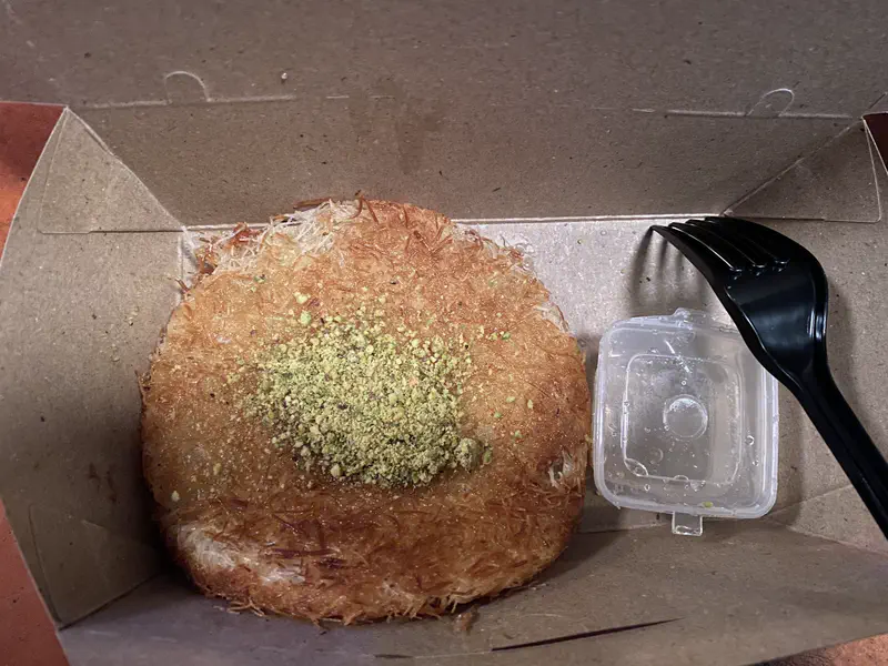 Box containing kunafa dessert topped with crushed pistachios and served with syrup.
