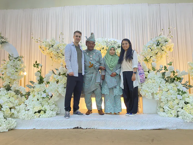 Bride and groom in traditional Malay attire posing with two guests on a decorated wedding stage with white flowers.