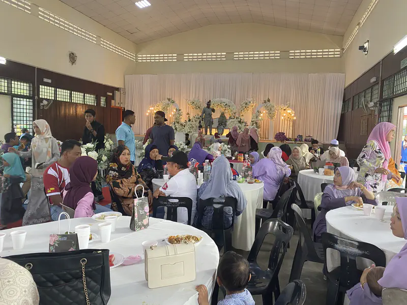 Guests seated at round tables inside a hall during a Malay wedding reception, with the bride and groom on stage.