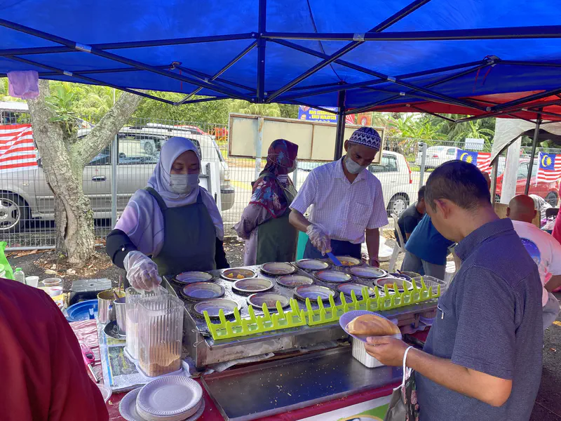 Street food stall vendors preparing apam balik pancakes on a griddle under a blue canopy.