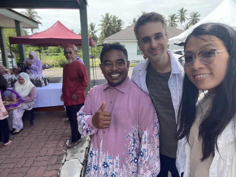 Three people smiling for a selfie at a Malay wedding event, with guests and a reception table in the background.