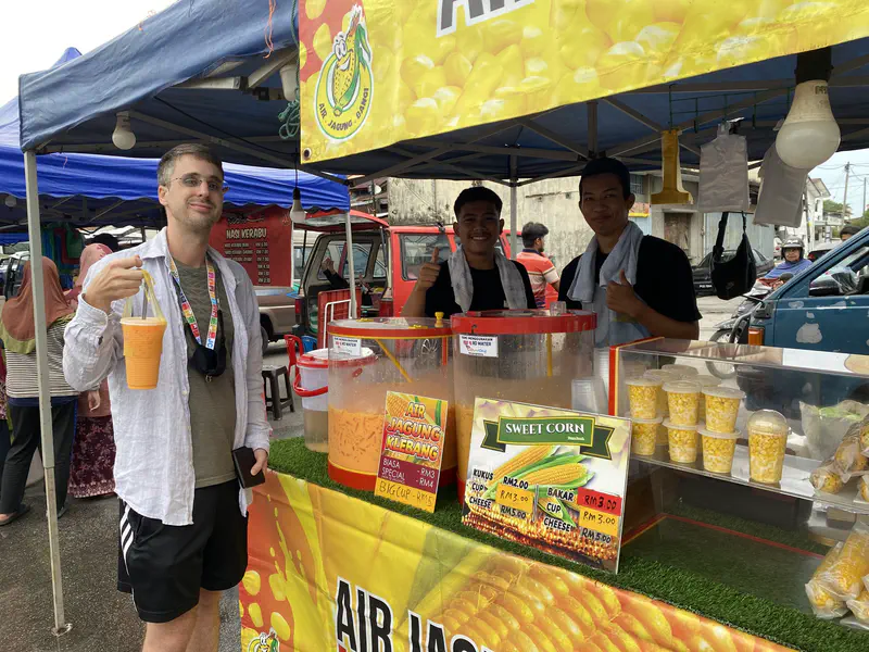Man holding a large cup of corn drink at a street stall with vendors selling air jagung and sweet corn.