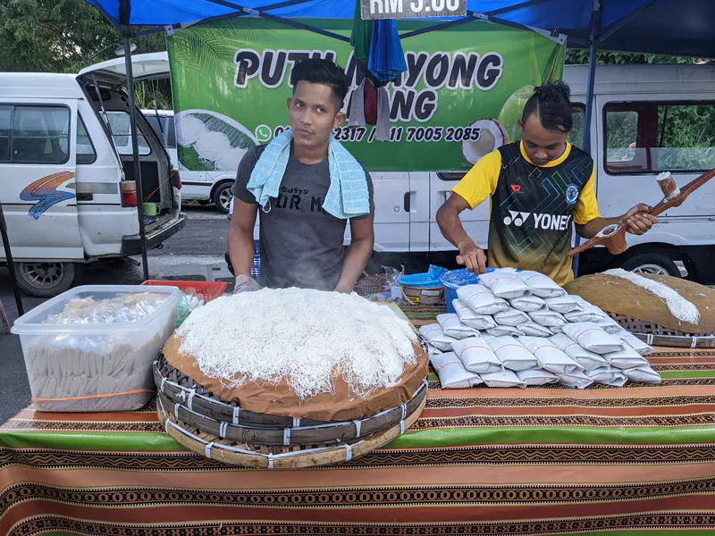 Street food stall selling putu mayong, with large trays of rice flour noodles and pre-packed portions on display.