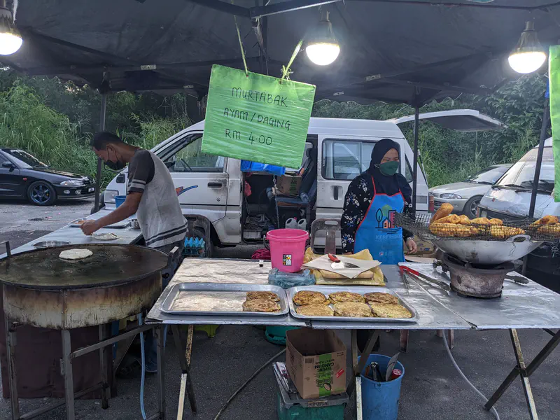 Street food stall with vendors preparing murtabak, a stuffed flatbread, on a large griddle with a sign reading 'Murtabak Ayam/Daging RM 4.00'.
