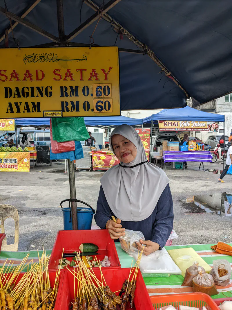 Woman vendor at a satay stall selling skewers of chicken and beef satay at a market.