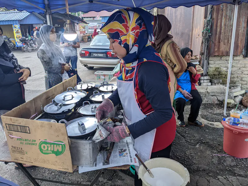 Vendor preparing apam balik or kuih at a market stall with several covered pans and customers waiting.