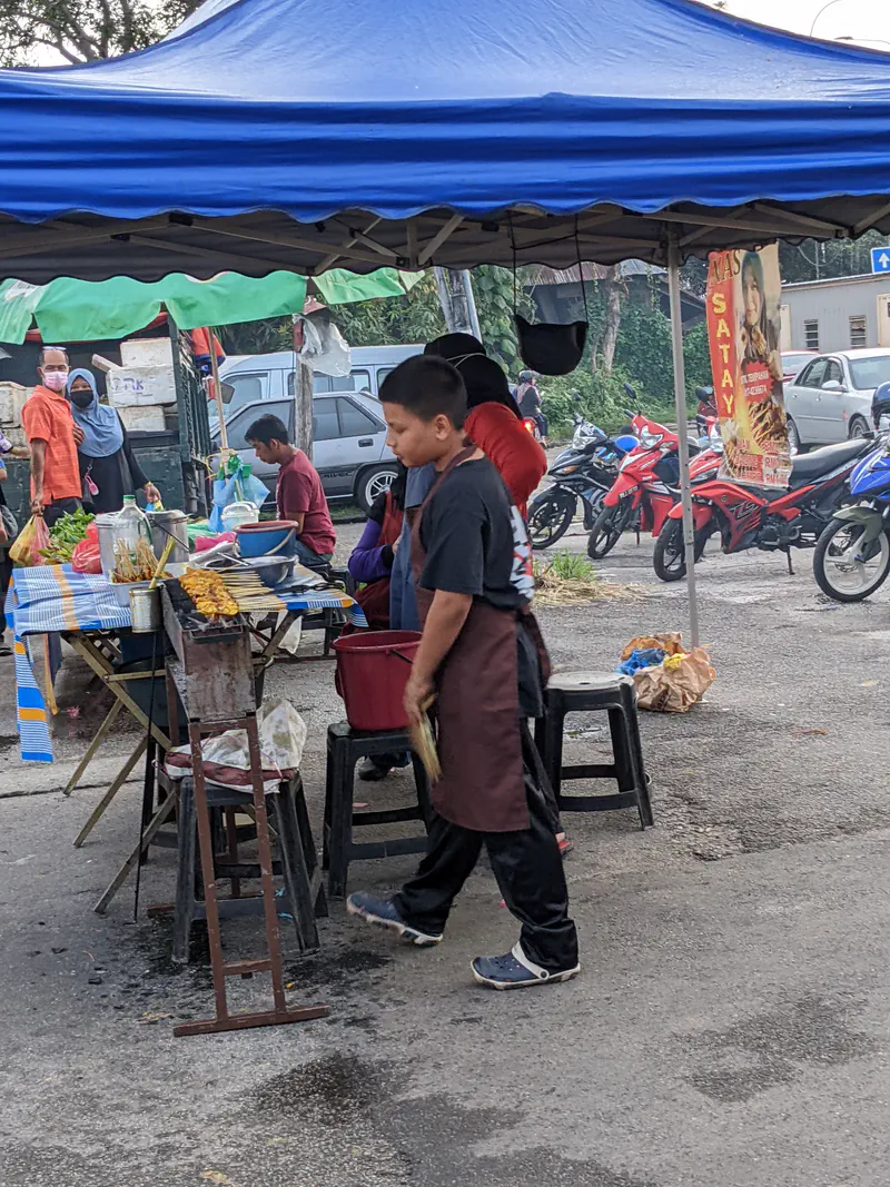 Young boy grilling satay skewers at a street food stall under a blue canopy.