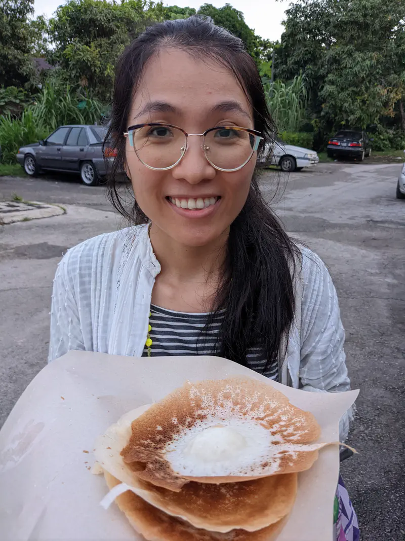 Woman smiling while holding freshly made apam balik on a paper sheet at an outdoor market.