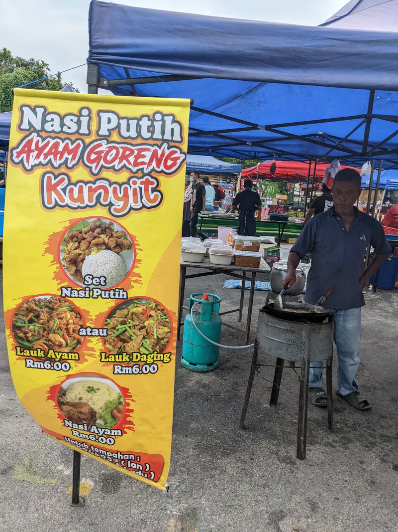 Vendor cooking nasi putih with ayam goreng kunyit (turmeric fried chicken) at a roadside stall with a bright yellow menu sign.