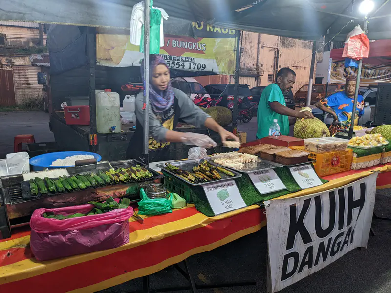 Night market stall selling traditional Malaysian snacks including grilled pulut panggang wrapped in banana leaves, kuih dangai, and fresh jackfruit.