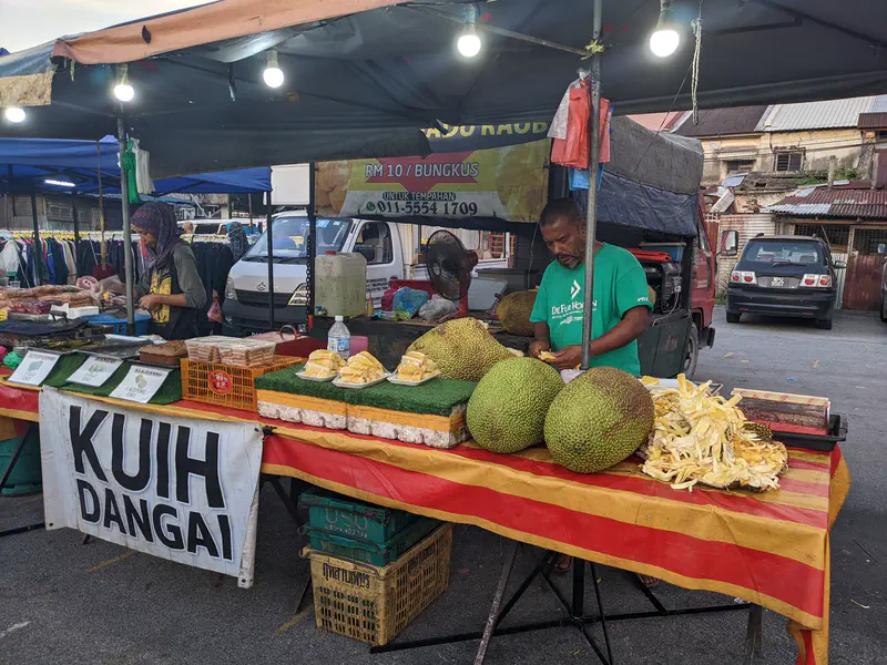 Market stall with vendors selling jackfruit and kuih dangai, with large jackfruits on display.