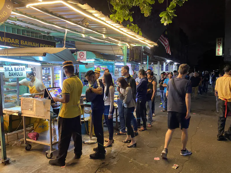 Crowd of people lining up at a popular nasi kandar street food stall at night, with bright lights illuminating the area.