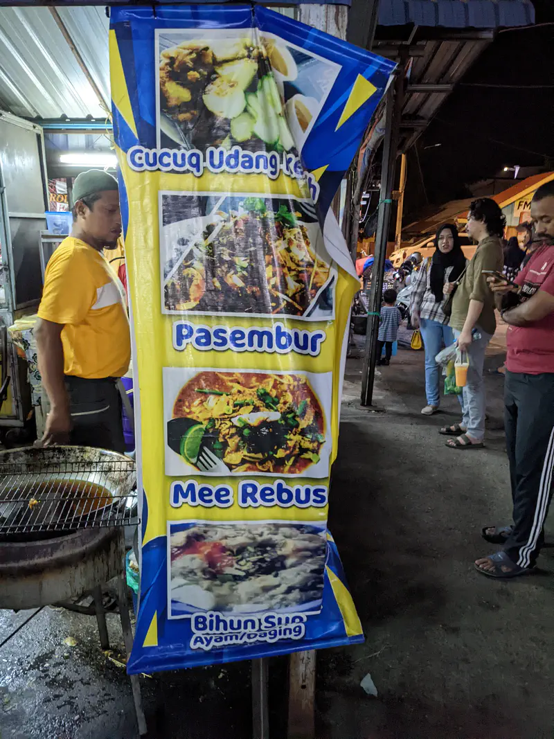 Food stall banner listing Malaysian dishes including cucuq udang, pasembur, mee rebus, and bihun sup, with customers nearby.