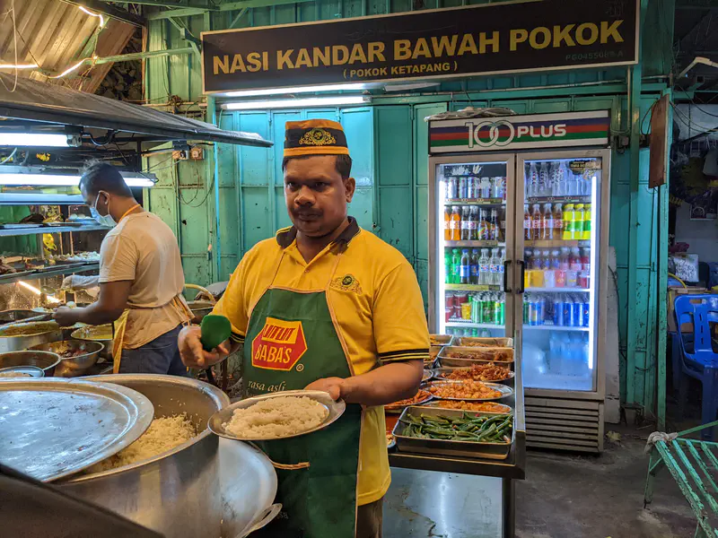 Vendor at Nasi Kandar Bawah Pokok stall serving rice with various side dishes displayed, inside a brightly lit shop.