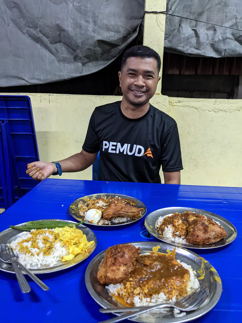 Smiling man sitting at a table with multiple plates of nasi kandar, rice topped with curry and fried chicken.