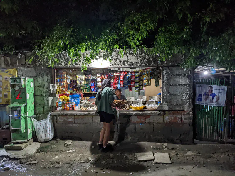 Small roadside store at night selling snacks, eggs, and goods with a customer buying items.