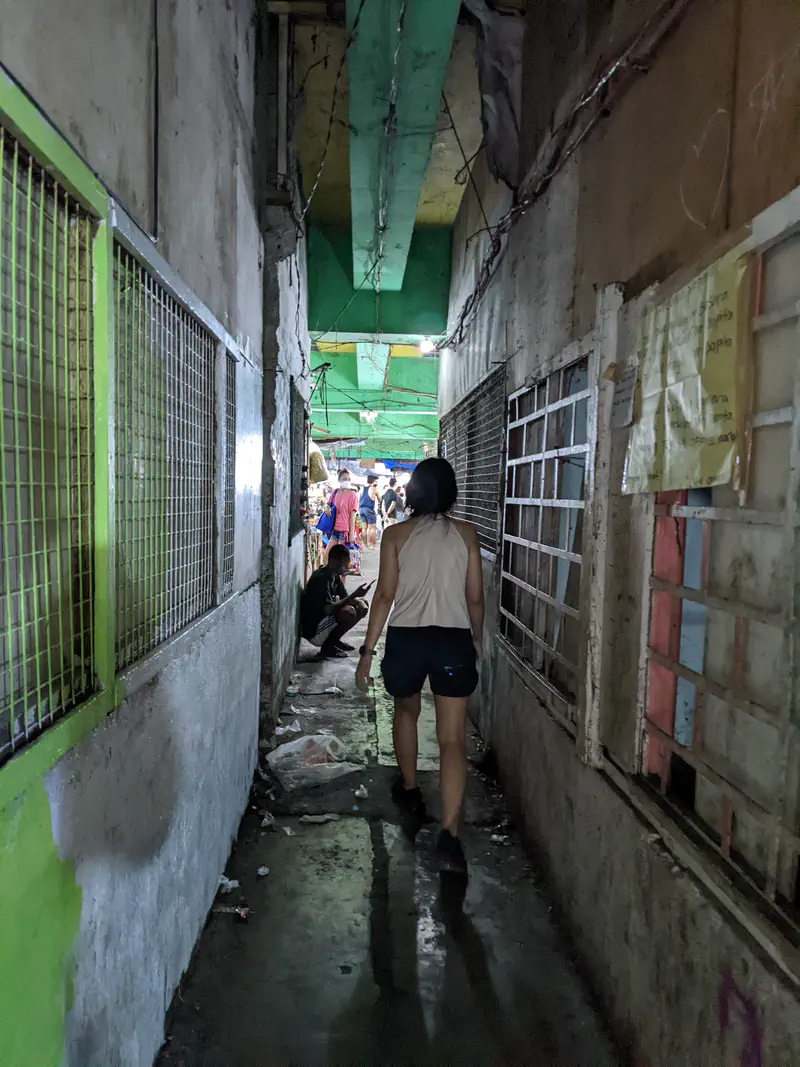 Narrow dim alleyway with a person walking toward a busy market area ahead.