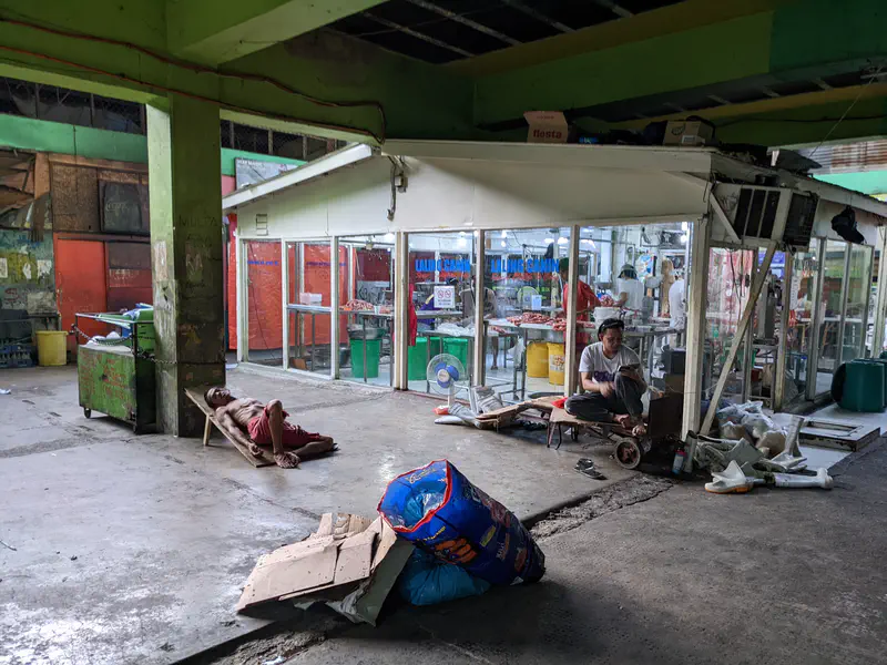 Two men resting outside a meat shop in a market, one lying on a cart and another sitting.