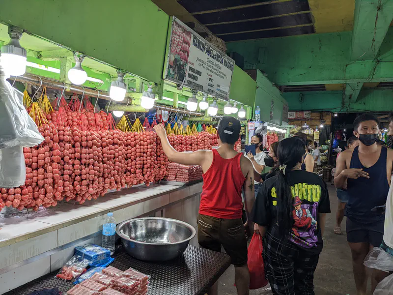 Market stall selling long strings of red sausages with several people shopping.