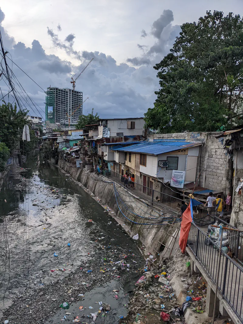 Polluted canal with floating trash beside informal houses and a high-rise under construction in the background.