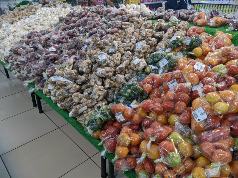 Assorted bagged vegetables including tomatoes, ginger, and sweet potatoes arranged on supermarket tables.