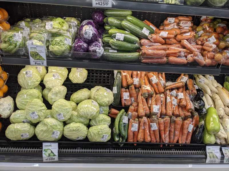 Supermarket display of wrapped cabbages, carrots, cucumbers, lettuce, and other vegetables on shelves.