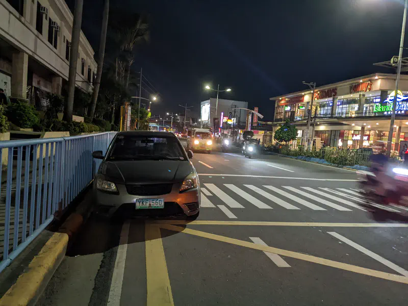 Car parked on a pedestrian lane beside a busy street at night with shops in the background.